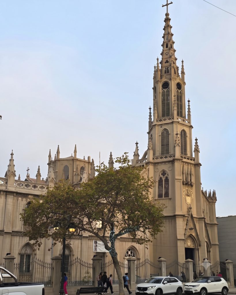 Iglesia de los Carmelitas de arquitectura neogótica de tonos beige con una alta torre central coronada por una cruz. En el frente se observa un árbol de copa verde, rejas de protección a la entrada, transeúntes caminando y vehículos estacionados.
