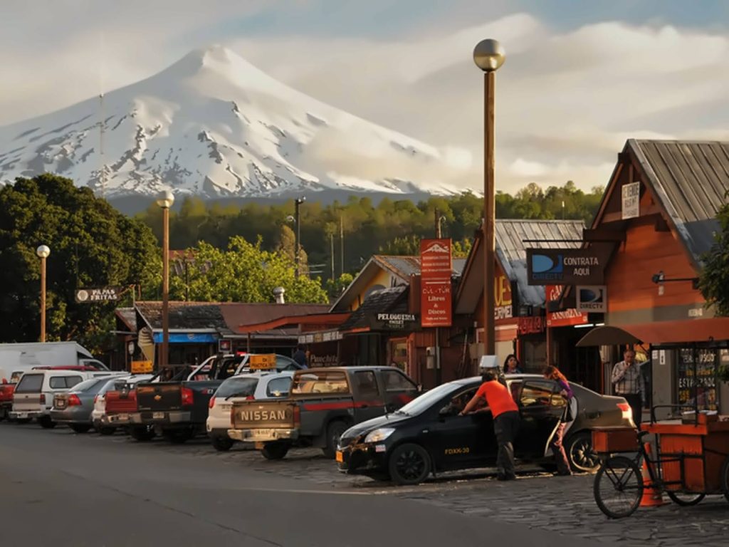 Calle del centro de Pucón con locales de madera y el volcán Villarrica nevado al fondo durante el atardecer.
