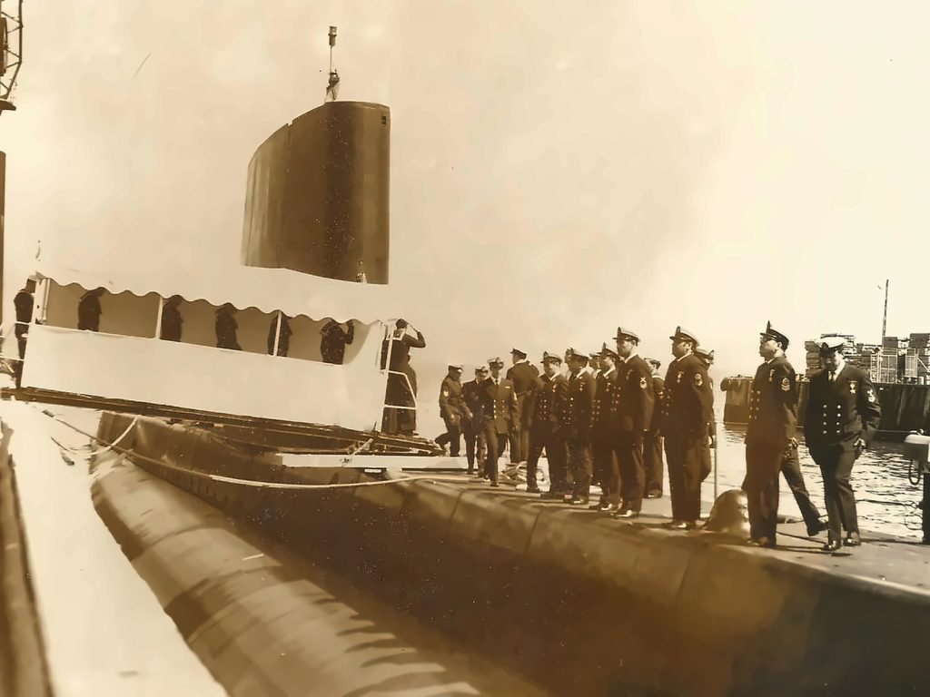Fotografía histórica en sepia de la tripulación de la Armada de Chile formada en la cubierta del Submarino O'Brien durante una ceremonia en el muelle.